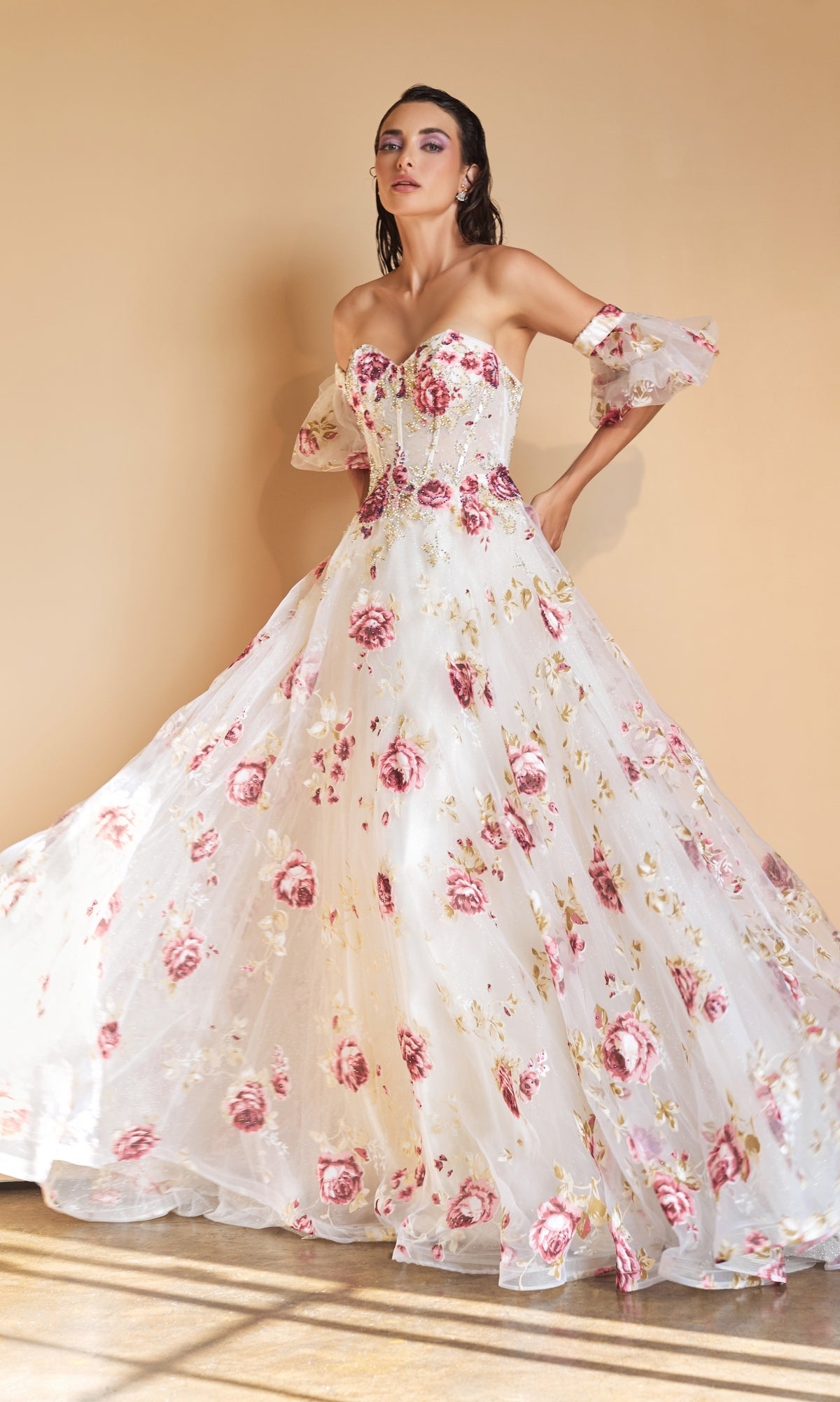 A woman models the Puff-Sleeve Strapless Floral-Print Ball Gown A1133, featuring sheer off-the-shoulder sleeves and pink floral patterns, creating an elegant look against a plain beige background.